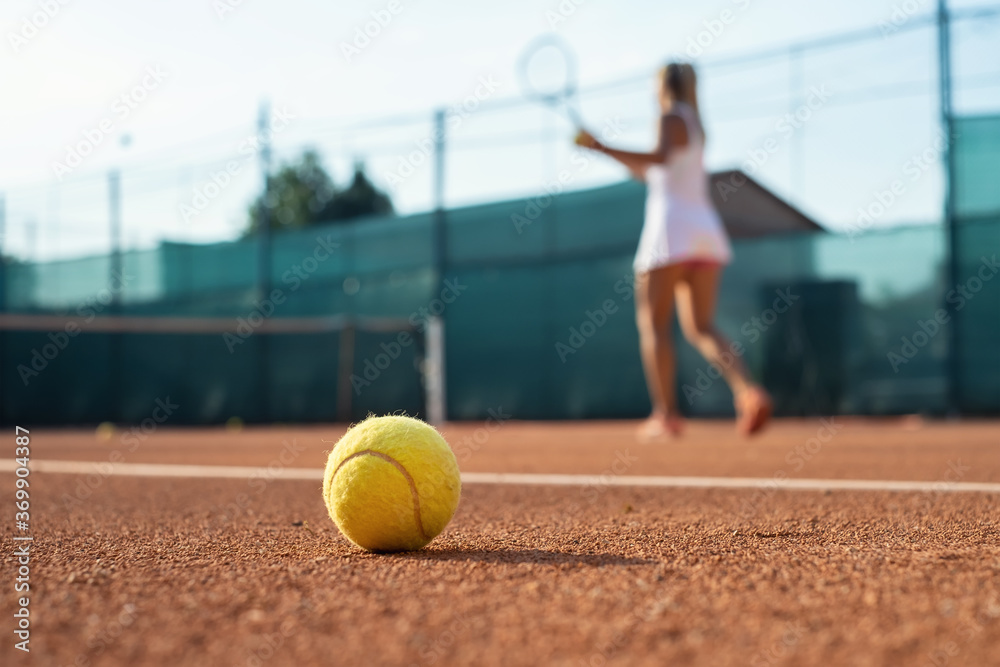 Healthy lifectyle. A young girl plays tennis on the court. The view on the ball and a tennis player. Dirt court. Sport background.