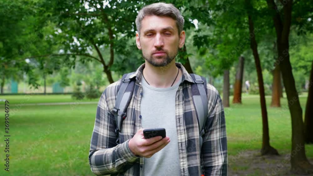 Gray-haired serious man tourist stands in park uses smartphone tap touch screen. Travel blogger guy in shirt, backpack holds smartphone in hand, raises head and looks camera. 4K slow motion footage Gray-haired serious man tourist stands in park uses smartphone tap touch screen. Travel blogger guy in shirt, backpack holds smartphone in hand, raises head and looks camera. 4K slow motion footage