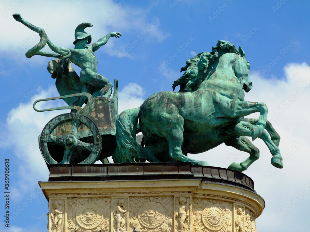 aged green statue of man with snake on a chariot. the symbol of war. Heroes' square in Budapest, Hungary.  summer light. blue sky. low angle view. travel and tourism concept. beautifully carved base.
