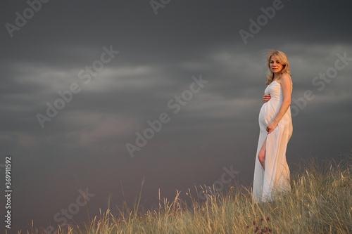 Pregnant young woman in a white dress on a posing standing on a mountain on a background of a dark cloudy sky