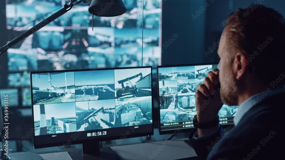 Male Officer Works on a Computer with Surveillance CCTV Video Footage in a Harbour Monitoring Center with Multiple Cameras on a Big Digital Screen. Employee Uses Radio to Give an Order or Report.