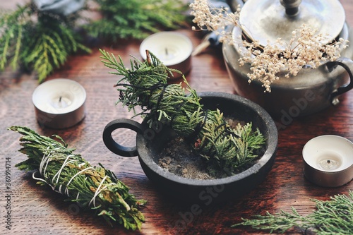 Cedar hand made smoke cleansing stick in cast iron ash holder burner on a wiccan witch altar. Cedar bundles for energy cleansing, alternative to sage smudge stick. Evergreens fir flowers in background