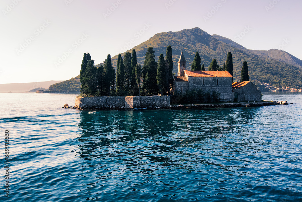 Catholic red roof medieval monastery, bell tower, masonry wall and cypress trees on an island in the Adriatic Sea. Little island in blue water of Kotor bay among Montenegro mountains. Island panorama