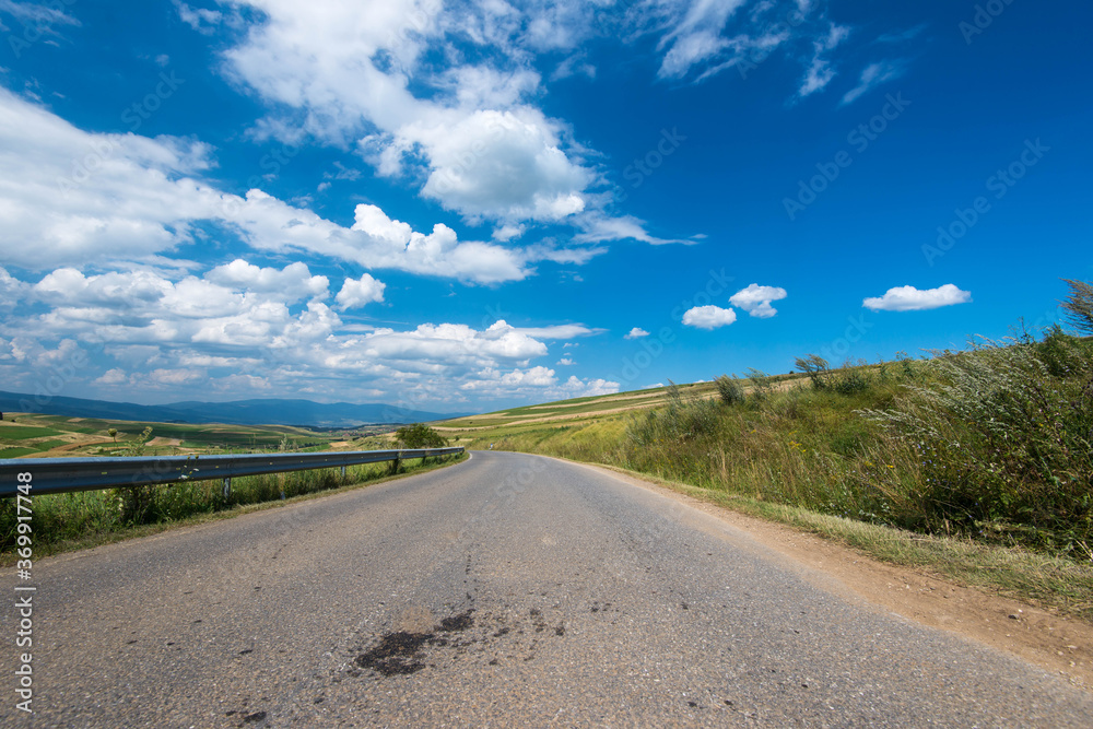 Asphalt road leading through green agricultural fields at summertime, beautiful blue sky with white clouds.