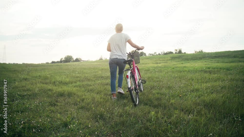 Cinematic shot of a young woman walking on a green field with a bicycle on sunset time. Cinematic shot of a young woman walking on a green field with a bicycle on sunset time.