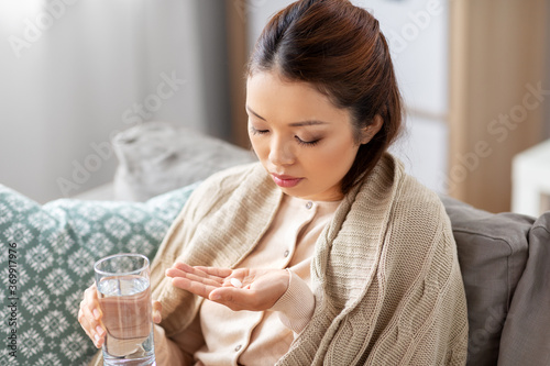 health, cold and people concept - sad sick young asian woman in blanket with medicine and glass of water at home