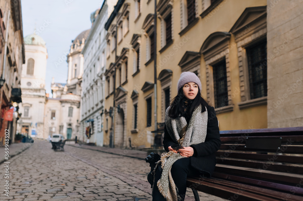 Young beautiful caucasian girl in autumn coat, scarf-wrap and angora hat sit on a street bench with smartphone in arm. Tourism concept. Ukraine Lviv. Young beautiful caucasian girl in autumn coat, scarf-wrap and angora hat sit on a street bench with smartphone in arm. Tourism concept. Ukraine Lviv.