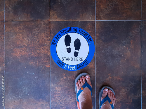 Round sticker with wording STAND HERE on floor and feet of person who stop stepping nearby the sticker. Sign to notice people keeping space from other. Social distabcing concept during Covid-19.
