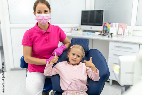 Lovely young kid in dental chair with dentist satisfied after repairing teeth. Small girl giving thumbs up to dentist.