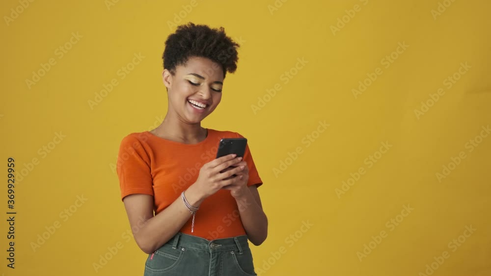 Satisfied attractive african woman playing on smartphone and having fun over yellow background