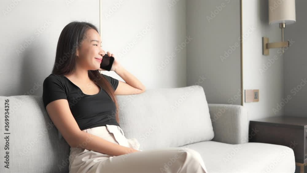 Happy young asian female talking on mobile phone while sitting on couch, slomo. Long distance communication concept, full frame Happy young asian female talking on mobile phone while sitting on couch, slomo. Long distance communication concept, full frame