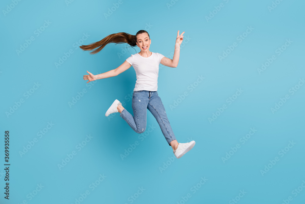 Full size photo of energetic excited crazy girl teen enjoy free time jump make v-sign wear white t-shirt gumshoes isolated over blue color background