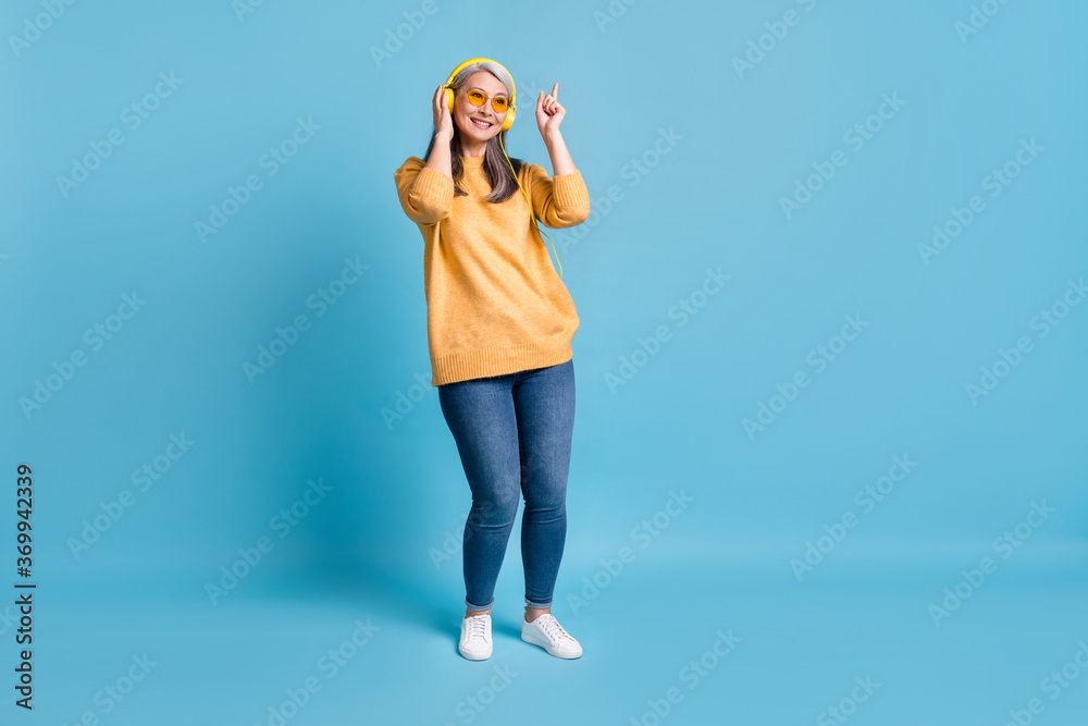 Full body size photo of lovely charming aged woman listen music hold headphones direct finger dancing alone home humming song wear sunglass yellow jumper vivid blue color background Full body size photo of lovely charming aged woman listen music hold headphones direct finger dancing alone home humming song wear sunglass yellow jumper vivid blue color background