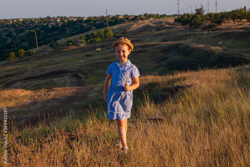 Adorable little girl in a straw hat and blue plaid summer dress in grass field with wild flowers in hands. Happy child with long blonde braided hair countryside landscape. Funny walking kid outdoor Adorable little girl in a straw hat and blue plaid summer dress in grass field with wild flowers in hands. Happy child with long blonde braided hair countryside landscape. Funny walking kid outdoor