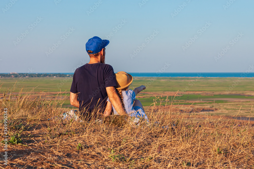 Father and daughter sitting by lakes on a nature lanscape. A little girl with long blond hair braid hugging with dad outdoor. Loving child embrace her daddy. Father's Day family greeting card faceless