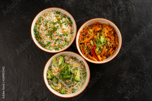 Three Asian side dishes in round bowls of rice, egg noodles, glass noodles and vegetables. Pan-Asian and Asian food and dishes on a stylish black concrete kitchen table. photo