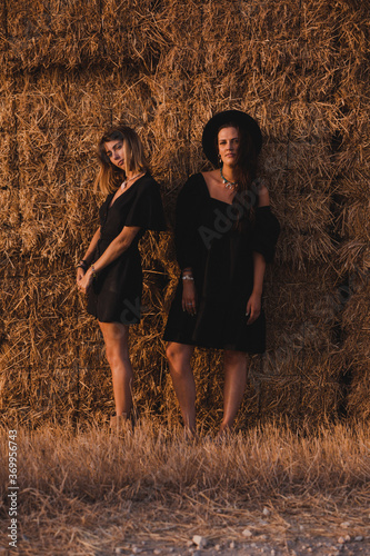 A couple of two young model women in a black dress in a straw bale background in summer in the golden hour of a summer evening