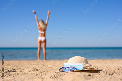 A straw hat, sunglasses and a medical mask lie on the beach by the sea. In the background, a girl stands with her hands up. Focus on the foreground. Vacation at sea during the coronavirus pandemic.