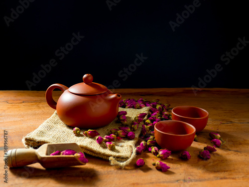 Teapot and teacup with rose tea leaves on wooden table and black background