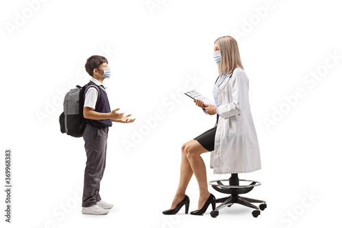 Schoolboy in a uniform with a face mask talking to a female doctor seated on a chair
