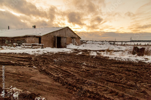 A barn in the middle of a snow-covered winter field next to a broken dirt road