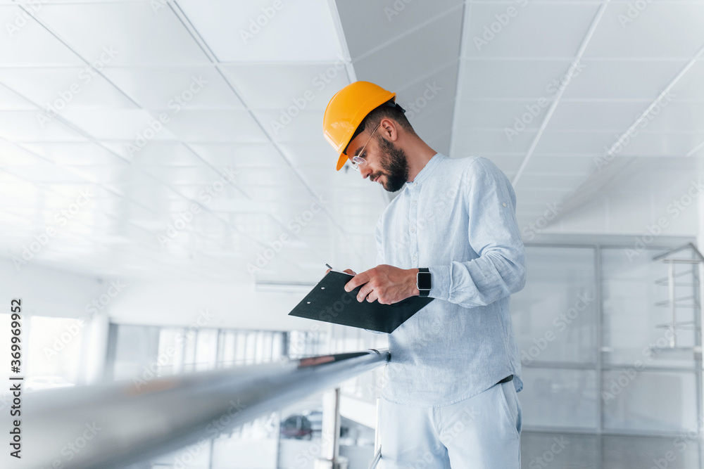 Holding notepad. Engineer in white clothes and orange protective hard hat standing and working indoors Holding notepad. Engineer in white clothes and orange protective hard hat standing and working indoors