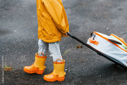 Close up view of kid in yellow waterproof cloak and boots walking outdoors with suitcase