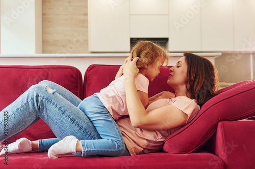 Lying down on red sofa. Young mother with her little daughter in casual clothes together indoors at home