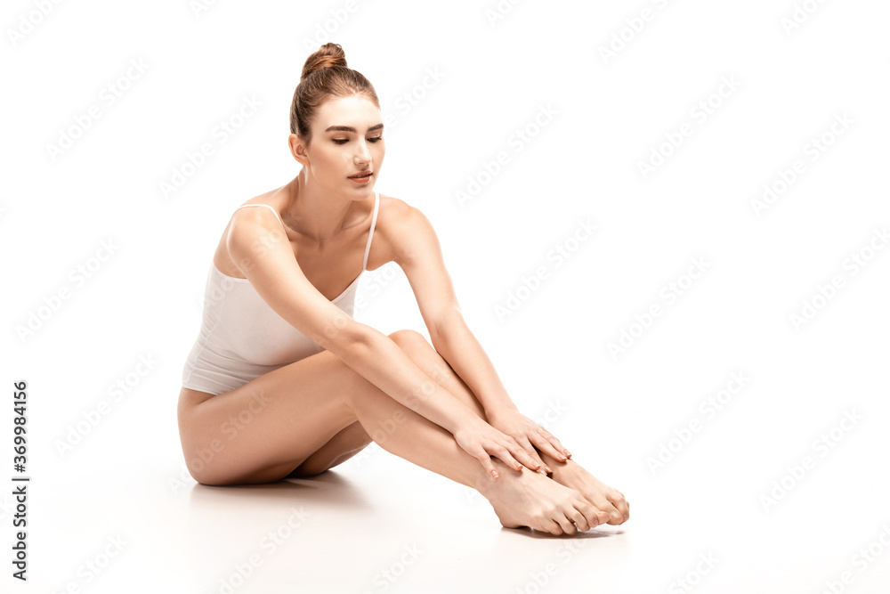 barefoot woman in bodysuit touching legs while sitting on white barefoot woman in bodysuit touching legs while sitting on white