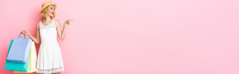 panoramic concept of woman in straw hat pointing with finger and holding shopping bags on pink