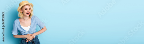panoramic crop of woman in straw hat laughing and touching belly on blue