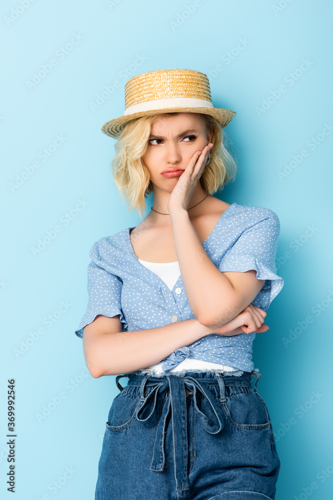 displeased woman in straw hat standing and touching face on blue displeased woman in straw hat standing and touching face on blue