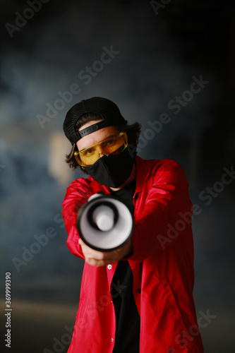 A young man of 25-30 years old in a black protective mask, yellow goggles, a cap and a red jacket holds a megaphone