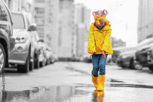 Adorable child girl in yellow raincoat and rubber boots in puddle on a parking, autumn walk. Childhood happiness concept