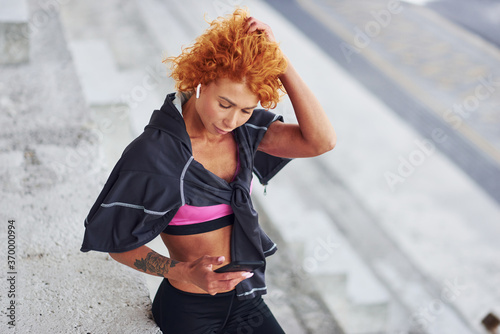 Young european redhead woman in sportive clothes standing on stairs outdoors with phone in hands