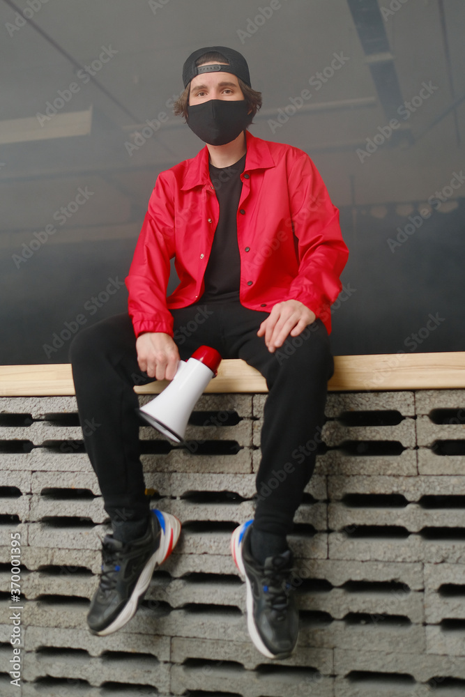 Emotional young man 25-30 years old in a black protective mask, a cap and a red jacket holding a megaphone against a dark background. Emotional young man 25-30 years old in a black protective mask, a cap and a red jacket holding a megaphone against a dark background.