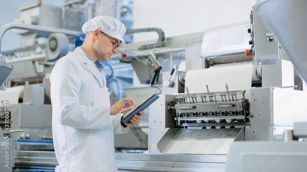 Young Male Quality Supervisor or Food Technician is Inspecting the Automated Production at a Dumpling Food Factory. Employee Uses a Tablet Computer for Work. He Wears White Sanitary Hat and Work Robe. Young Male Quality Supervisor or Food Technician is Inspecting the Automated Production at a Dumpling Food Factory. Employee Uses a Tablet Computer for Work. He Wears White Sanitary Hat and Work Robe.
