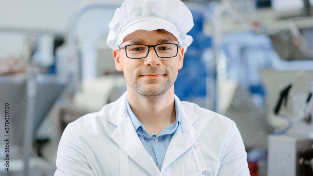 Young Male Quality Supervisor or Technician in Glasses is Working on a Food Factory. Employee Looks and Smiles at Camera. He Wears a White Sanitary Hat and Work Robe.