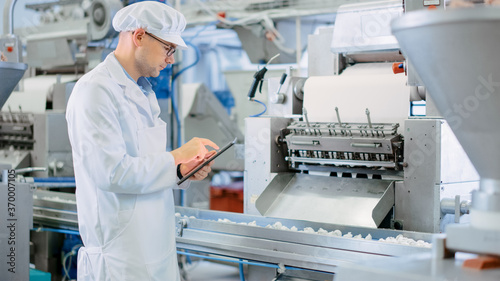 Young Male Quality Supervisor or Food Technician is Inspecting the Automated Production at a Dumpling Factory. Employee Uses a Tablet Computer for Work. He Wears a White Sanitary Hat and Work Robe.