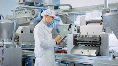 Young Male Quality Supervisor or Food Technician is Inspecting the Automated Production at a Dumpling Food Factory. Employee Uses a Tablet Computer for Work. He Wears White Sanitary Hat and Work Robe.