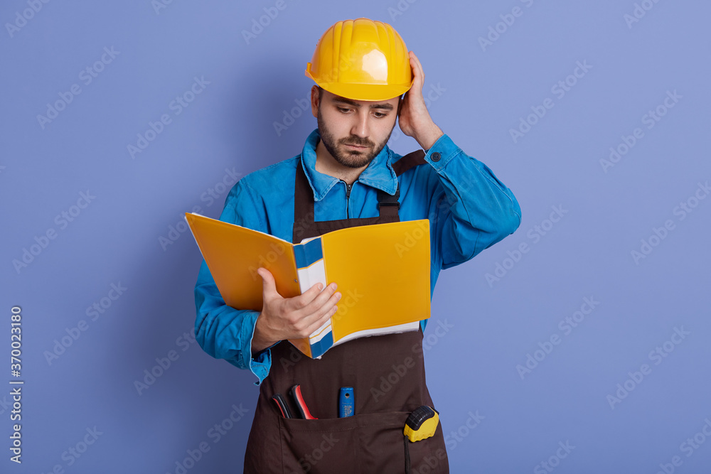 Construction engineer hold yellow paper folder read information about new project, do not understand details, looks at documents with puzzled facial expression. Construction engineer hold yellow paper folder read information about new project, do not understand details, looks at documents with puzzled facial expression.