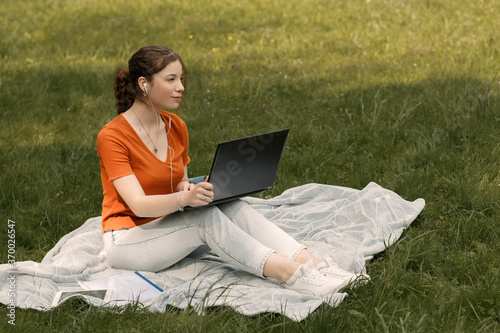 Cheerful girl working with laptop outside. Young student in the park with copy space. Woman sitting on the plaid. Full length portrait with gadgets.