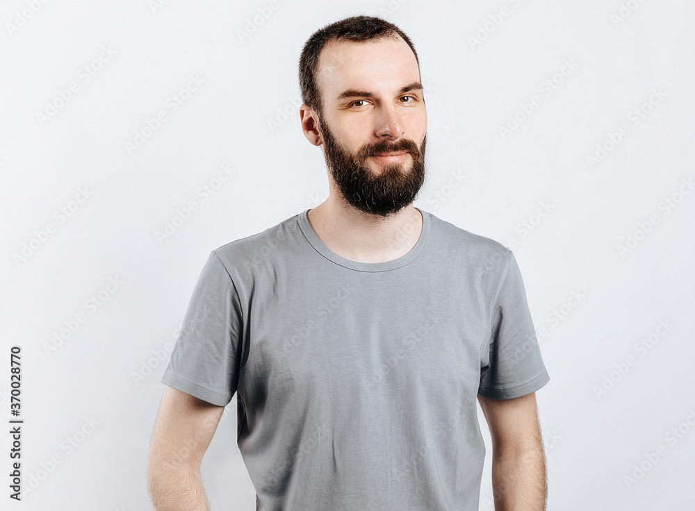 A handsome brutal brunette man with a beard in a gray shirt smiles and looks at the camera. Place for advertising on a gray background. A handsome brutal brunette man with a beard in a gray shirt smiles and looks at the camera. Place for advertising on a gray background.
