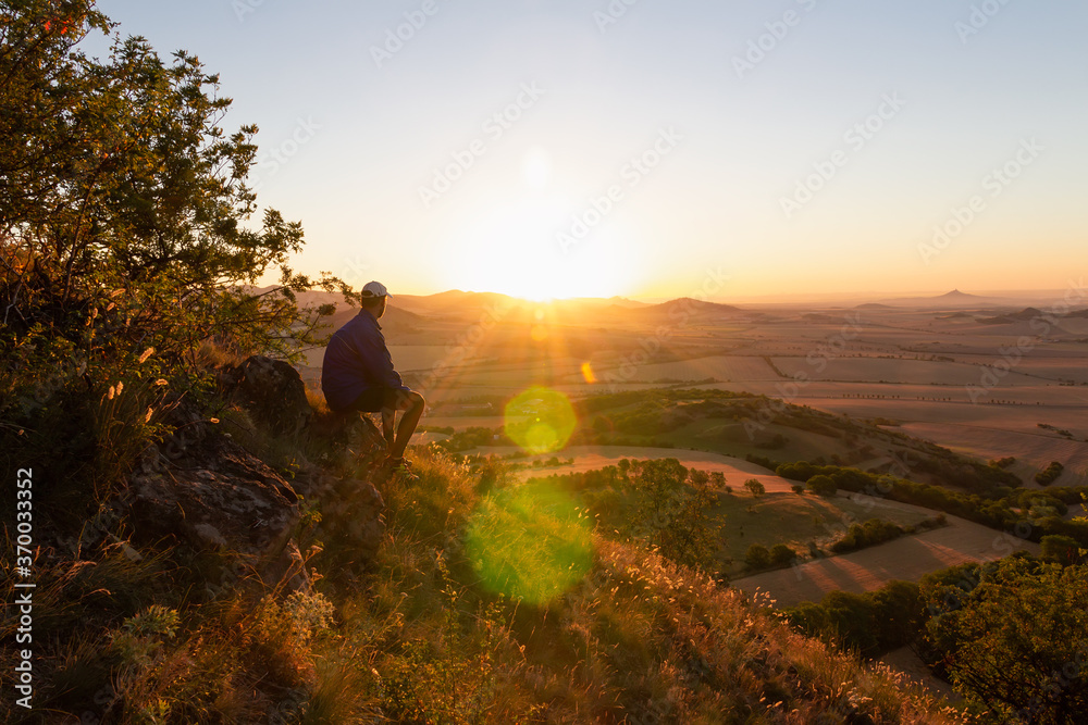 Young man with backpack sitting on hill and looking to Czech ore mountain valley at sunrise. Czech landscape