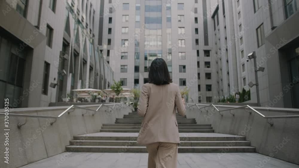 Shot back woman wearing formal suit walking hold coffee and tablet computer look around outside business cellphone city smartphone slow motion Shot back woman wearing formal suit walking hold coffee and tablet computer look around outside business cellphone city smartphone slow motion