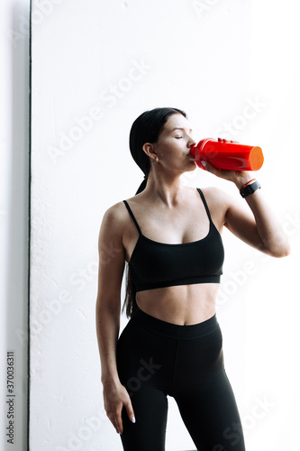 Brunette in sportswear drinks protein food from red shaker on white background