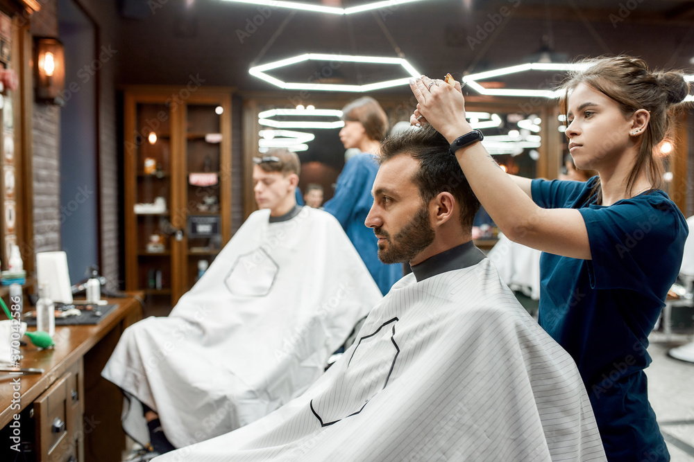 Side view of a barber girl making new trendy haircut for a young handsome bearded man sitting in barbershop chair. Visiting hair salon Side view of a barber girl making new trendy haircut for a young handsome bearded man sitting in barbershop chair. Visiting hair salon