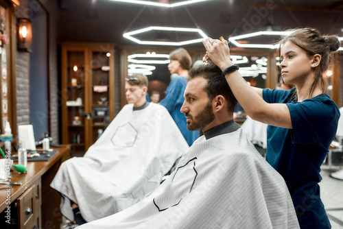 Side view of a barber girl making new trendy haircut for a young handsome bearded man sitting in barbershop chair. Visiting hair salon