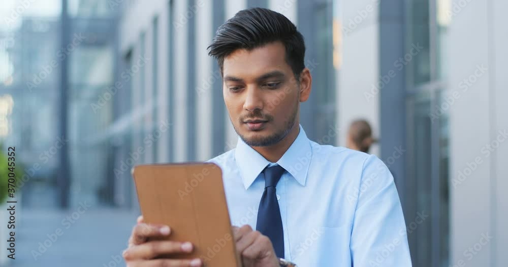 Handsome Hindu young businessman in tie using tablet device at street. Man from India in business style tapping and scrolling on gadget computer. Outdoors. Close up. Handsome Hindu young businessman in tie using tablet device at street. Man from India in business style tapping and scrolling on gadget computer. Outdoors. Close up.