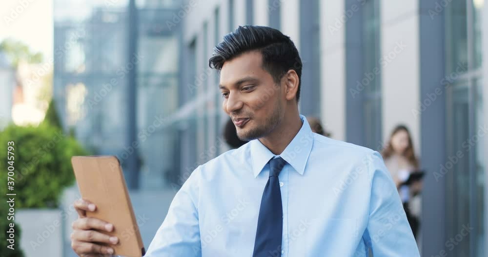 Hindu handsome young businessman in tie standing at street at business center and talking via videochat on smartphone. Man from India videochatting on webcam of mobile phone. Video call. Hindu handsome young businessman in tie standing at street at business center and talking via videochat on smartphone. Man from India videochatting on webcam of mobile phone. Video call.